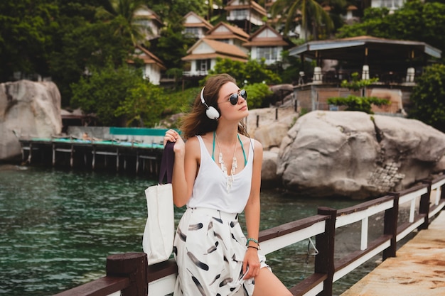 Jonge stijlvolle vrouw permanent op pier, wandelen, luisteren naar muziek op koptelefoon, zomerkleding, witte rok, handtas, azuurblauw water, landschapsachtergrond, tropische lagune, vakantie, reizen in Azië