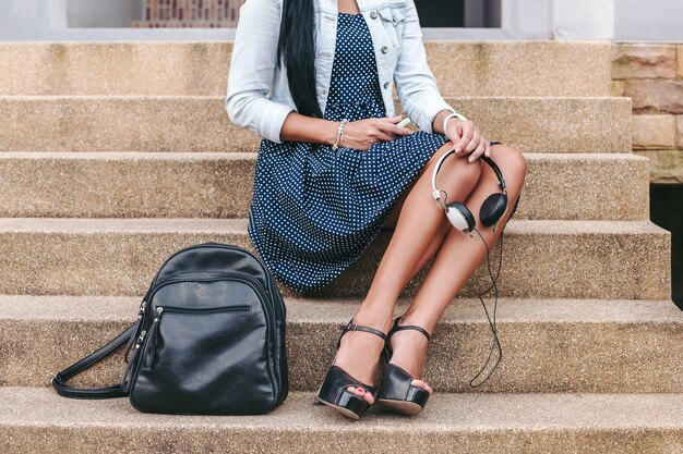 Jonge stijlvolle vrouw zittend op de trap, koptelefoon in handen, rugzak, lange benen, schoenen met hoge hakken, gebruinde huid, close-up details, accessoires