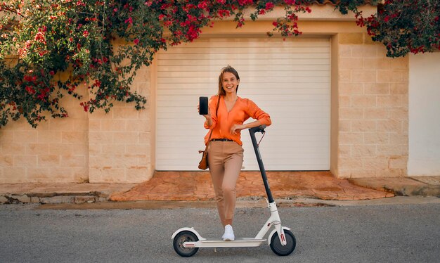 Jonge stijlvolle zakenvrouw of vrouwelijke student met een elektrische scooter toont het smartphonescherm terwijl ze op straat staat in een stad met bloemen.