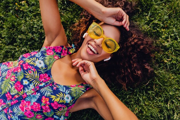 Jonge stijlvolle zwarte vrouw luisteren naar muziek op draadloze oortelefoons met plezier in park, zomer fashion stijl, kleurrijke hipster outfit, liggend op het gras, van bovenaf bekijken