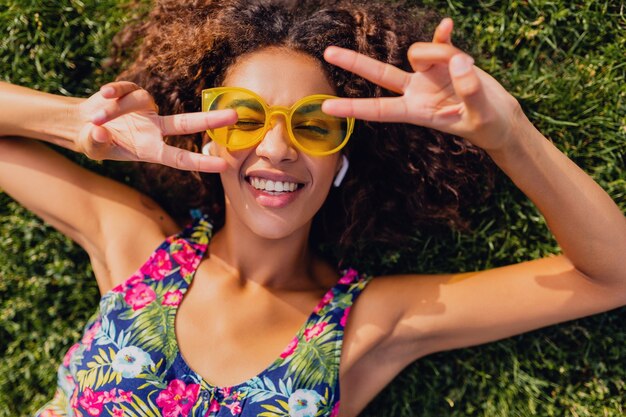 Jonge stijlvolle zwarte vrouw luisteren naar muziek op draadloze oortelefoons plezier liggend op gras in park, zomer fashion stijl, kleurrijke hipster outfit, van bovenaf bekijken