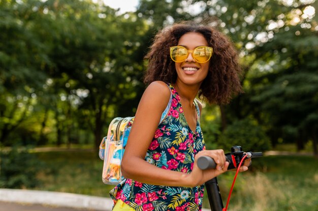 Jonge stijlvolle zwarte vrouw plezier in park rijden op elektrische kick scooter in zomer fashion stijl, kleurrijke hipster outfit, rugzak en gele zonnebril dragen