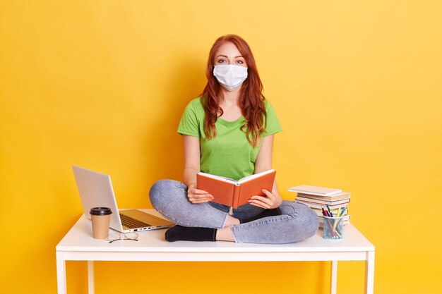 Jonge student in medische masker thuis studeren in quarantaine, verveeld van afstandsonderwijs, zittend met gekruiste benen op witte tafel met boek in handen.