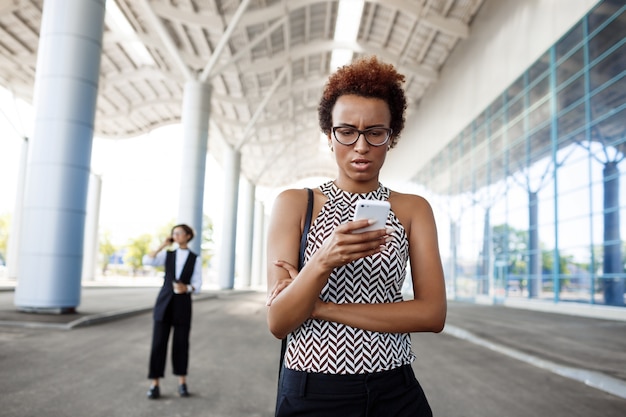 Jonge succesvolle Afrikaanse onderneemster die telefoon over commercieel centrum bekijkt.