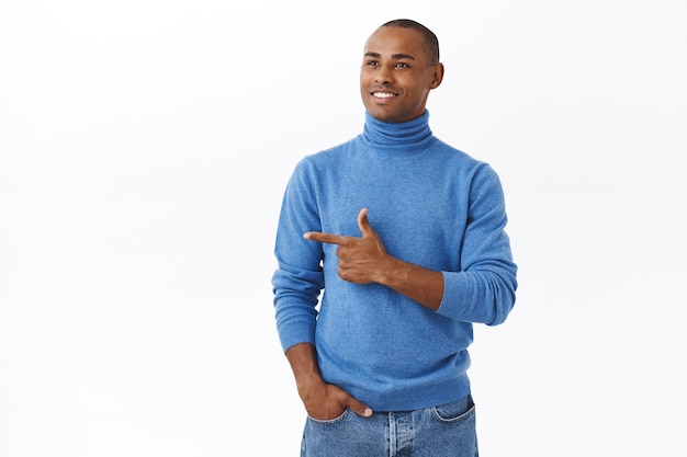 Jonge succesvolle Afro-Amerikaanse man, gekleed in een blauwe coltrui, kijkt met een tevreden gezicht en wijst naar links