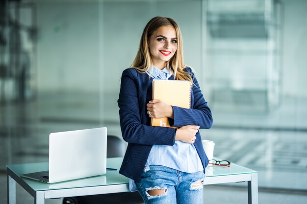 Jonge succesvolle vrouw in casual kleding bedrijf notebook werk staande in de buurt van wit bureau met laptop in kantoor. Prestatie carrière bedrijfsconcept.