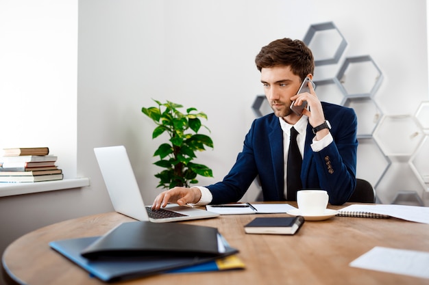 Jonge succesvolle zakenman die op telefoon, bureauachtergrond spreekt.