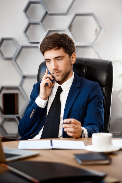 Jonge succesvolle zakenman die op telefoon, bureauachtergrond spreekt.