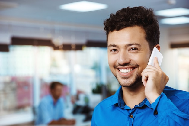 Jonge succesvolle zakenman die op telefoon spreekt, die over bureau glimlacht