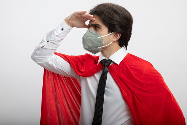 Jonge superheld kerel die stropdas en medisch masker draagt ?? die afstand met hand bekijkt die op wit wordt geïsoleerd
