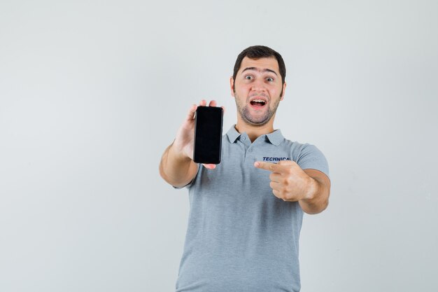 Jonge technicus in grijs uniform wijzend op mobiele telefoon en vrolijk, vooraanzicht kijken.