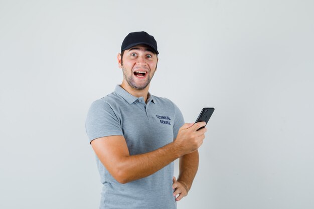 Jonge technicus leest de berichten op zijn telefoon en glimlacht terwijl hij zijn hand op de taille houdt in een grijs uniform en er verbaasd uitziet.