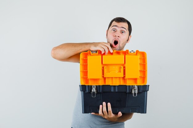 Jonge technicus toolbox openen met zijn beide handen in grijs uniform en versuft op zoek.