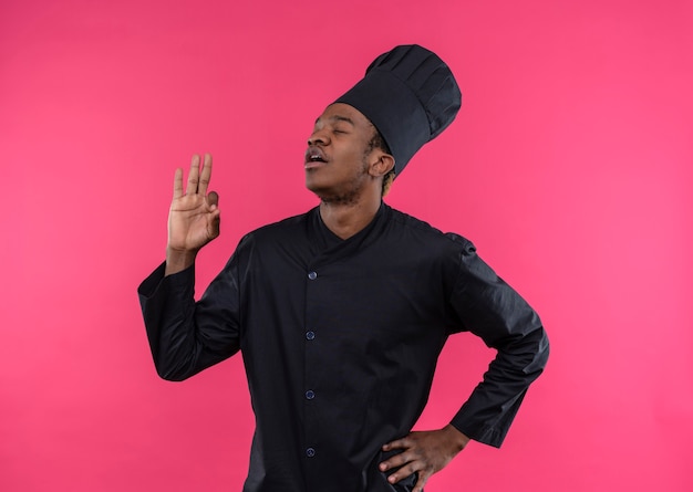 Jonge tevreden Afro-Amerikaanse kok in chef-kok uniforme gebaren ok handteken geïsoleerd op roze muur