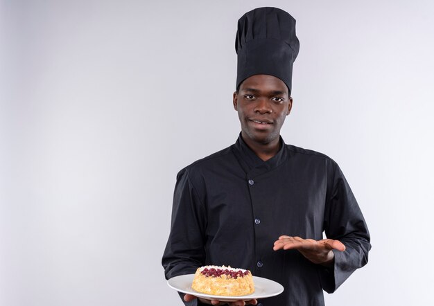 Jonge tevreden Afro-Amerikaanse kok in uniform chef houdt cake op plaat en punten met hand op wit met kopie ruimte