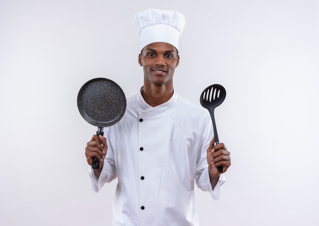 Jonge tevreden Afro-Amerikaanse kok in uniform chef houdt koekenpan en spatel op geïsoleerde witte muur