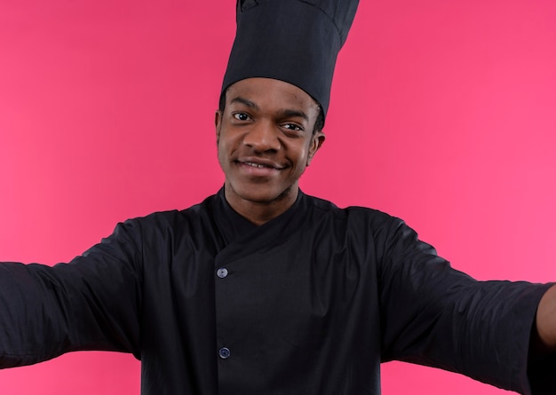 Jonge tevreden Afro-Amerikaanse kok in uniform chef-kok beweert te knuffelen geïsoleerd op roze muur