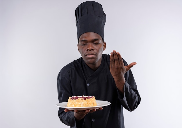 Jonge tevreden afro-amerikaanse kok in uniform chef-kok houdt en beweert cake op plaat op wit met kopie ruimte te ruiken