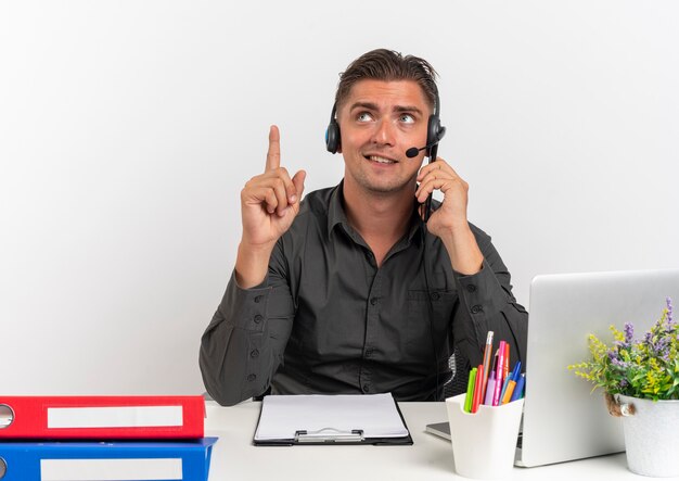 Jonge tevreden blonde kantoormedewerker man op koptelefoon zit aan bureau met office-hulpprogramma's met behulp van laptop kijkt en wijst omhoog geïsoleerd op een witte achtergrond met kopie ruimte