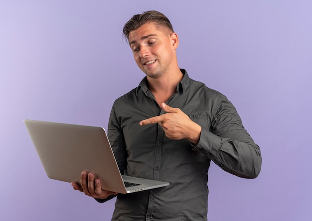 Jonge tevreden blonde knappe man houdt en wijst op laptop geïsoleerd op violette achtergrond