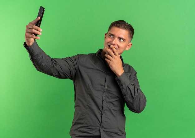 Jonge tevreden blonde knappe man kijkt naar telefoon selfie te nemen en legt de hand op de mond geïsoleerd op groene ruimte met kopie ruimte