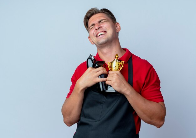 Jonge tevreden blonde mannelijke kapper in uniform houdt kapper tools en winnaar beker met gesloten ogen geïsoleerd op een witte ruimte met kopie ruimte