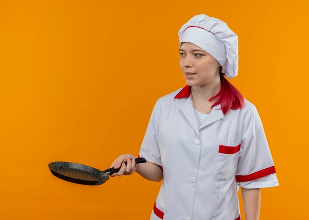 Jonge tevreden blonde vrouwelijke chef-kok in eenvormige chef-kok houdt koekenpan en kijkt naar kant geïsoleerd op oranje muur