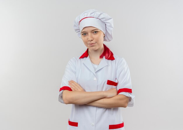 Jonge tevreden blonde vrouwelijke chef-kok in eenvormige chef-kok kruist armen en kijkt geïsoleerd op een witte muur