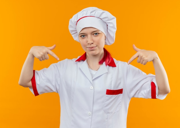 Jonge tevreden blonde vrouwelijke chef-kok in eenvormige chef-kok wijst naar beneden met beide handen geïsoleerd op oranje muur