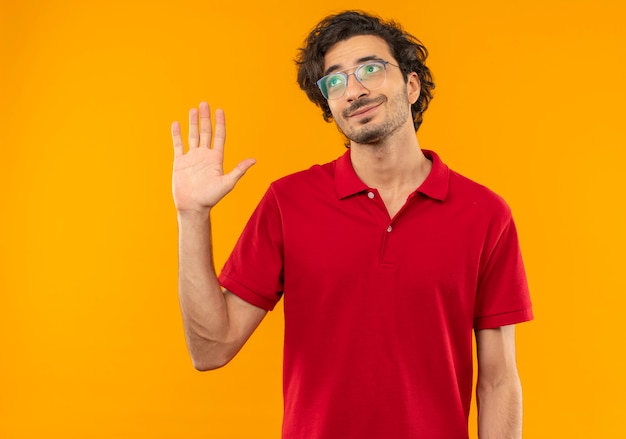 Jonge tevreden man in rood shirt met optische bril steekt hand omhoog en kijkt omhoog geïsoleerd op oranje muur