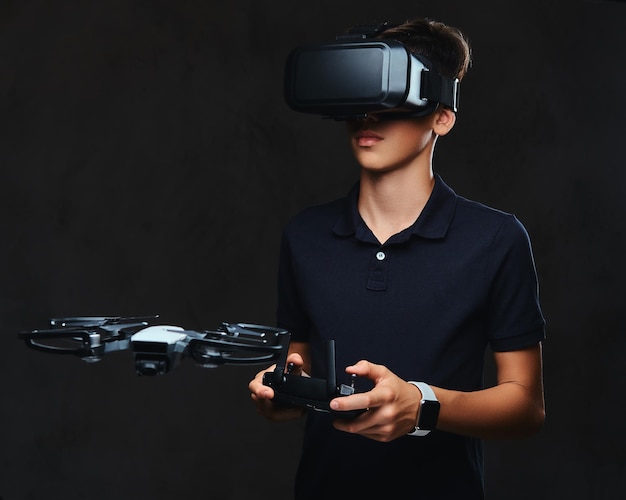 Jonge tiener gekleed in een zwart t-shirt draagt een virtual reality-bril en bestuurt de quadcopter met behulp van de afstandsbediening. Geïsoleerd op een donkere achtergrond.