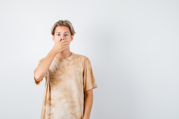 Jonge tienerjongen die de hand op de mond houdt in een t-shirt en er geagiteerd uitziet. vooraanzicht.