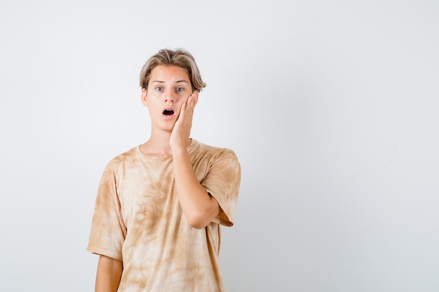 Jonge tienerjongen die de hand op de wang houdt in een t-shirt en er verrast uitziet, vooraanzicht.