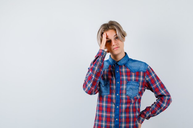 Jonge tienerjongen die lijdt aan hoofdpijn in een geruit overhemd en er verdrietig uitziet. vooraanzicht.