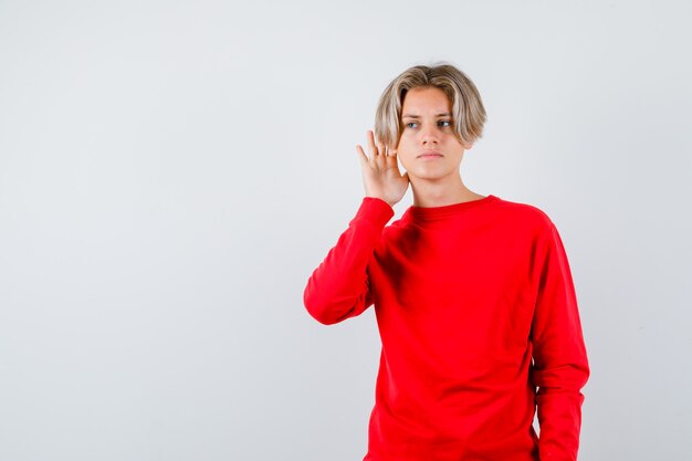 Jonge tienerjongen die privégesprek in rode sweater afluistert en verward kijkt. vooraanzicht.