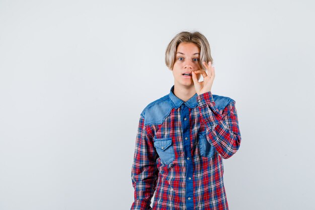 Jonge tienerjongen die zip-gebaar in geruit overhemd toont en voorzichtig kijkt
