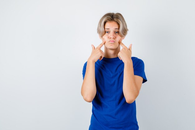 Jonge tienerjongen in blauw t-shirt die vingers op de wangen drukt en er somber uitziet, vooraanzicht.