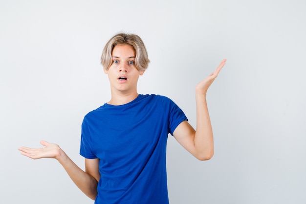 Jonge tienerjongen in blauw t-shirt met hulpeloos gebaar en verbaasd, vooraanzicht.