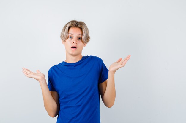 Jonge tienerjongen in blauw t-shirt met hulpeloos gebaar en verbijsterd, vooraanzicht.