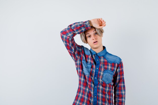 Jonge tienerjongen in geruit overhemd die de hand op het hoofd houdt en er bedroefd uitziet, vooraanzicht.