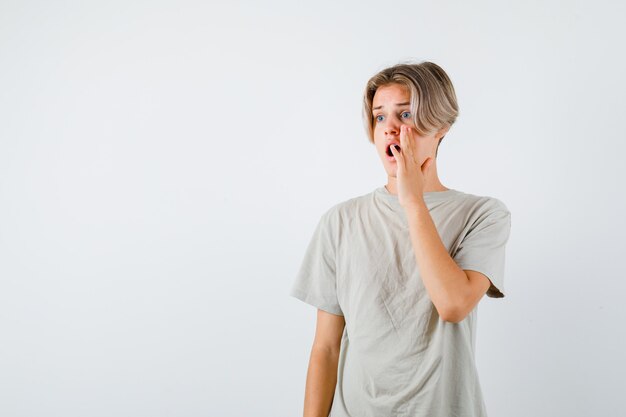 Jonge tienerjongen in t-shirt die hand in de buurt van open mond houdt terwijl hij wegkijkt en geschokt kijkt, vooraanzicht.