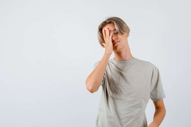 Jonge tienerjongen in t-shirt die zijn hand op zijn gezicht houdt en er vermoeid uitziet