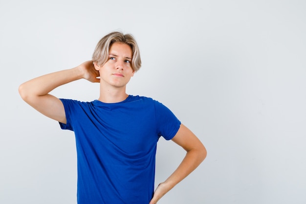 Jonge tienerjongen met hand achter hoofd, omhoog kijkend in blauw t-shirt en peinzend kijkend, vooraanzicht.