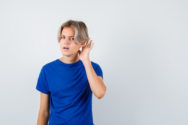 Jonge tienerjongen met hand achter oor in blauw t-shirt en verward kijkend. vooraanzicht.