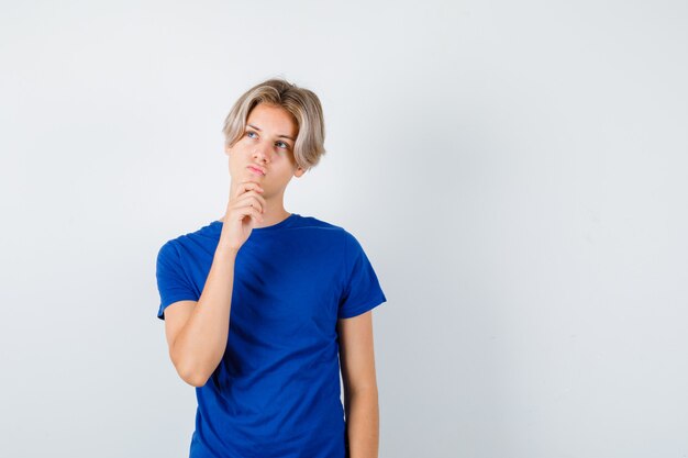 Jonge tienerjongen met hand op kin, omhoog kijkend in blauw t-shirt en nadenkend, vooraanzicht.