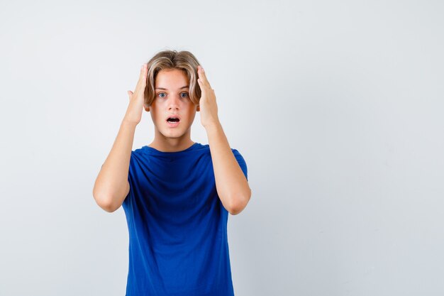 Jonge tienerjongen met handen op hoofd in blauw t-shirt en geagiteerd, vooraanzicht.