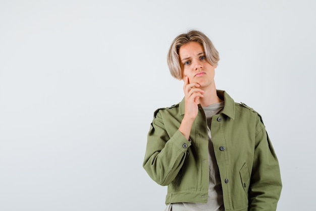 Jonge tienerjongen met vinger op wang in groen jasje en somber kijkend. vooraanzicht.