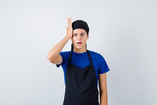 Jonge tienerkok met hand op hoofd in t-shirt, schort en weemoedig, vooraanzicht.