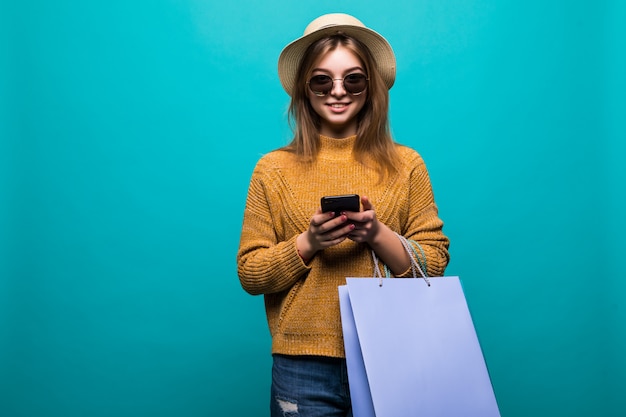 Jonge tienervrouw in zonnebril en hoed die iets in smartphone kijken en het winkelen zakken in haar handen houden