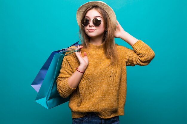 Jonge tienervrouw in zonnebril en hoedenholding het winkelen zakken in haar handen die zo geluk voelen dat op groene muur wordt geïsoleerd